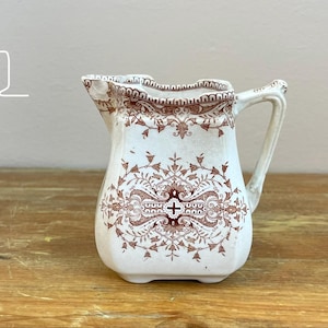 May include: A white ceramic creamer pitcher featuring a brown transferware pattern with intricate floral and scrollwork motifs, a scalloped rim, and a sturdy handle. The body displays a central cross symbol surrounded by detailed decorative elements.