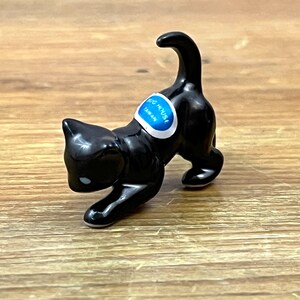 May include: A small, glossy black cat figurine in a playful pouncing pose with its tail held high. The cat features a white and blue oval sticker on its back with the text BUG HOUSE TAIWAN printed in white letters. The figurine has a smooth finish.