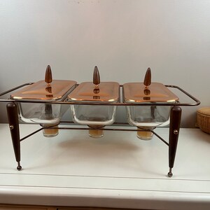 May include: A three-section copper and glass serving dish with wooden handles sits on a metal stand with wooden legs.