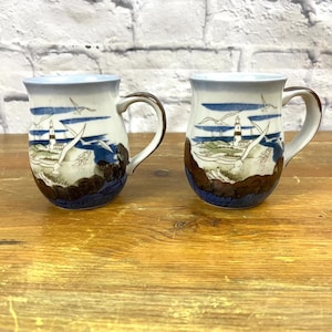 May include: Two ceramic mugs with a lighthouse and seagull design. The mugs are white with blue and brown accents.