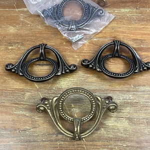 May include: An assortment of antique-style drawer pulls in bronze and brass tones. The pulls feature a ring design with decorative detailing and are shown on a wooden surface. One pull is still in its original packaging.