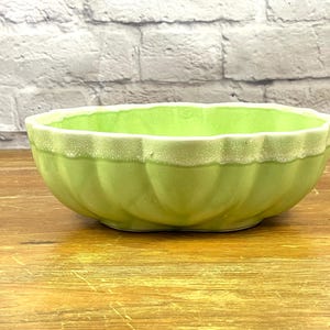 May include: A light green ceramic bowl with a scalloped rim and a ribbed exterior. The rim has a textured, off-white band. The bowl is set on a wooden surface against a white brick wall.
