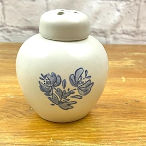 May include: A small, round, off-white ceramic jar with a matching lid featuring a blue hand-painted floral design on the front. The jar has a smooth, matte finish and a rounded body shape, with a lid that includes small holes on the top surface.