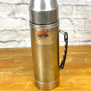 May include: A silver flask with a black handle. The flask has a label that reads "America Uni-Vac" in red letters.