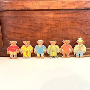 May include: Seven paper cutouts of teddy bears wearing different outfits. The bears are all brown and have a yellow face. The outfits include a red and white striped shirt, a yellow robe, a blue uniform, a green uniform, a red shirt, and a white shirt with a black vest and a top hat.