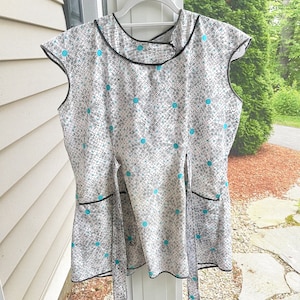 May include: A white apron with a black and blue geometric pattern. The apron has a tie at the waist and two pockets.