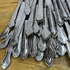 May include: A pile of silver-plated butter knives with ornate floral designs. The knives are arranged in a fan shape, with the handles pointing towards the top of the image.