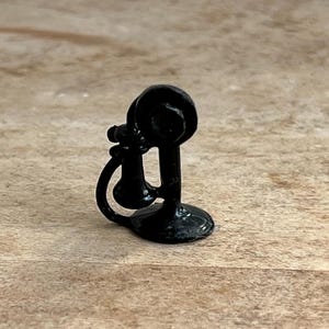 May include: A small, black vintage telephone miniature with a circular earpiece and mouthpiece, connected by a curved handle. The phone rests on a round base. The miniature is made of a shiny, dark material.