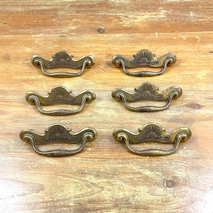May include: Six antique brass drawer pulls with ornate floral designs. The pulls have a curved shape and a center ring.