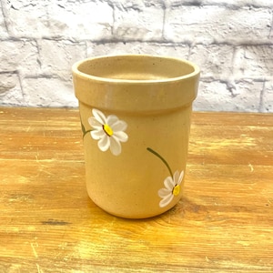 May include: A tan ceramic pot with hand-painted white daisies and yellow centers. The pot is cylindrical with a slightly flared rim. The daisies have green stems and leaves. The pot is likely used for planting flowers or herbs.