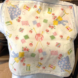 Puede incluir: Babero de tela blanca con borde azul claro con un diseño temático de costura. El diseño incluye carretes de hilo, tijeras, motivos florales y una bola de lana con agujas de tejer. El babero tiene una regla impresa a lo largo del borde.