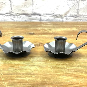 May include: Two antique-style, dark gray metal candle holders. Each has a scalloped base, a central cup for the candle, and a curved handle. The candle holders are displayed on a wooden surface.