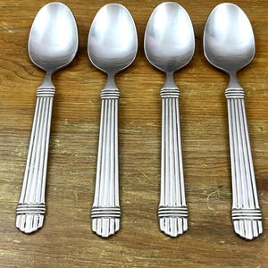 May include: Four silver spoons with an elegant design. Each spoon has an oval bowl and a handle decorated with vertical lines and bands. They are arranged on a wooden surface.