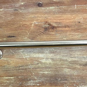 May include: A polished chrome towel bar with rounded ends and decorative finials. The bar is mounted to a wooden surface.