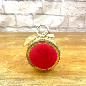 May include: A vintage pin cushion with a gold-toned metal base and a vibrant red velvet top. The cushion is adorned with a decorative trim and a gold ribbon, and features a small metal ring for hanging.