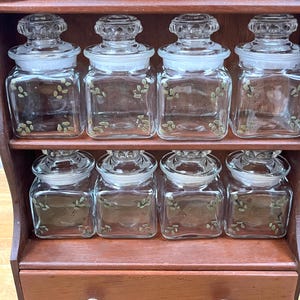 May include: A vintage wooden spice rack with eight clear glass jars. Each jar has a square shape, a glass lid, and a decorative leaf pattern. The rack is a dark brown color.