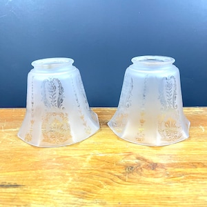 May include: Two frosted glass lampshades with a gold floral design. The lampshades are white and have a scalloped edge.