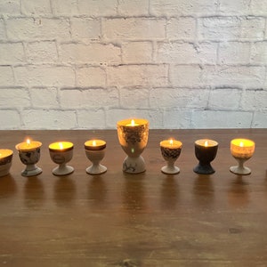 Może przedstawiać: Dziewięć białych ceramicznych świeczników z zapalonymi świeczkami. Świeczniki mają różne wzory i projekty. Świeczniki są ułożone w rzędzie na drewnianej powierzchni.