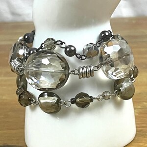 May include: A layered bracelet featuring faceted, clear, and smoky gray beads. The bracelet has a mix of round and irregularly shaped beads, strung on a silver-toned chain. The bracelet is displayed on a white surface.