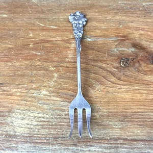 May include: A silver-colored fork with a floral design on the handle. The fork has three tines and a detailed flower and leaf pattern at the top. The background is a wooden surface.