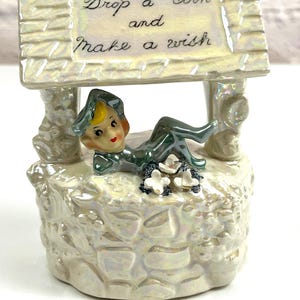 Pode incluir: Uma figura de poço dos desejos de cerâmica com um duende verde sentado na borda. O poço tem uma placa que diz "Drop a coin and make a wish".