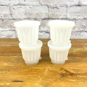 May include: Four white, fluted plastic containers with lids are stacked in pairs on a wooden surface. The containers have a classic design and are likely for food storage or portioning. The background features a white brick wall.