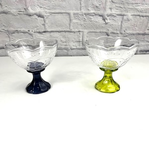 Puede incluir: Dos cuencos de postre de vidrio transparente con bordes festoneados y un patrón decorativo. Los cuencos están sobre pedestales, uno azul y otro verde.
