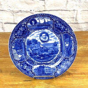 Puede incluir: Plato de cerámica azul y blanco con un diseño decorativo que representa una escena del desembarco de Hendrick Hudson. El plato tiene un borde floral azul y blanco e incluye el texto "Landing of Hendrick Hudson".