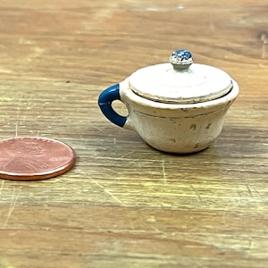 May include: A miniature white ceramic tea cup with a lid and a blue handle. The lid is topped with a blue and white decorative knob. A penny is next to the tea cup for size comparison. The tea cup is on a wooden surface.