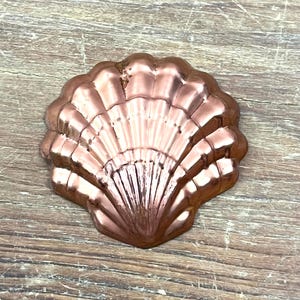May include: A metallic copper-colored scallop shell mold with a shiny, reflective surface. The object features a fan-shaped design with distinct, raised vertical ridges radiating from the base to the scalloped outer edge of the shell.