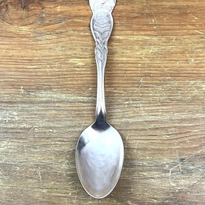 Könnte beinhalten: Ein silberner Löffel mit einem verzierten Griff mit einem Wappenmuster. Die Löffelschale ist oval und reflektiert das Licht. Der Griff hat detaillierte Gravuren. Der Löffel ist wahrscheinlich Vintage oder antik.