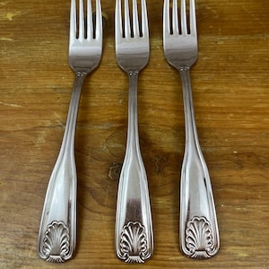 May include: Three silver forks with a shell design on the handles are arranged on a wooden surface. The forks have tines and a decorative pattern at the base of the handle. The forks are made of a shiny metal.