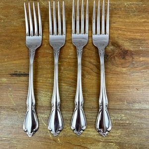 May include: Four silver forks with ornate handles are arranged on a wooden surface. The forks have a classic design with a decorative pattern on the handle and tines. The forks are made of a shiny, reflective metal.