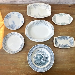 May include: A collection of antique white and blue porcelain plates and serving dishes. The plates feature floral and scenic designs. The arrangement includes round plates, oval platters, and rectangular serving dishes, all with delicate patterns.