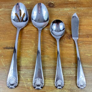 May include: A set of four silver-colored serving utensils, including a slotted spoon, a serving spoon, a small spoon, and a butter knife. Each piece has a decorative shell design at the base of the handle. The utensils are arranged on a wooden surface.
