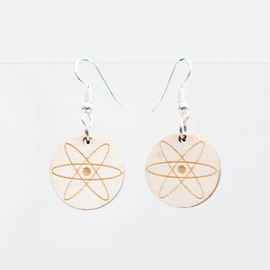 Könnte beinhalten: Ein Paar Ohrringe aus Holz mit einem silbernen Haken. Jeder Ohrring hat ein kreisförmiges Design mit einem lasergravierten Atom-Symbol.