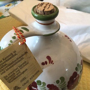 May include: White ceramic pitcher with a green and red floral design. The pitcher has a cork stopper and a tag that reads "Unsere Keramik ist spülmaschinenfest, backfest, mikrowellenherdbeständig und lebensmittelecht. Sie erwerben hiermit Keramik von höchster Qualität und Wertbeständigkeit. IHRE GRAF KERAMIK"