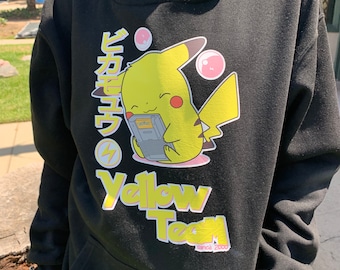 May include: Black hoodie featuring a yellow Pikachu graphic with Japanese text and the words "Yellow Team" in yellow. The design includes pink bubbles and a lightning bolt. The hoodie has a front pocket.