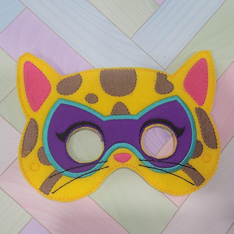 May include: A yellow felt cat mask featuring brown spots, pink inner ears, and a pink nose. The mask includes a purple superhero-style eye mask overlay outlined in teal, with black embroidered eyelashes and whiskers on the yellow base.