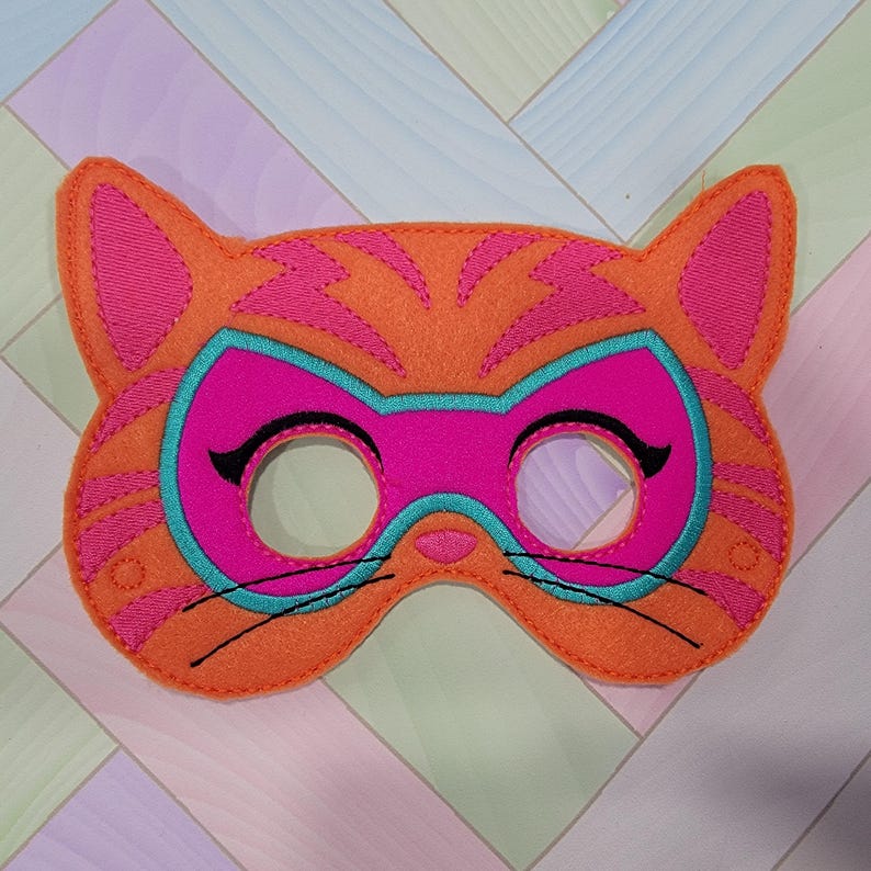 May include: An orange felt cat mask featuring pink tiger stripes, a bright pink eye mask area outlined in teal, and black embroidered whiskers and eyelashes. The mask has two circular eye cutouts and is constructed with visible stitching.