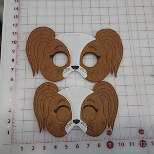May include: Two brown and white felt dog masks featuring long ears, stitched eye openings, and embroidered black details including eyebrows, eyelashes, a nose, and whiskers. The masks are positioned on a cutting mat with centimetre markings.