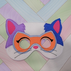 May include: A felt cat mask featuring a white base with a purple left ear and a blue right ear. The mask includes an orange eye patch area, pink inner ears, a small pink nose, and black embroidered eyelashes and whiskers around the eye openings.