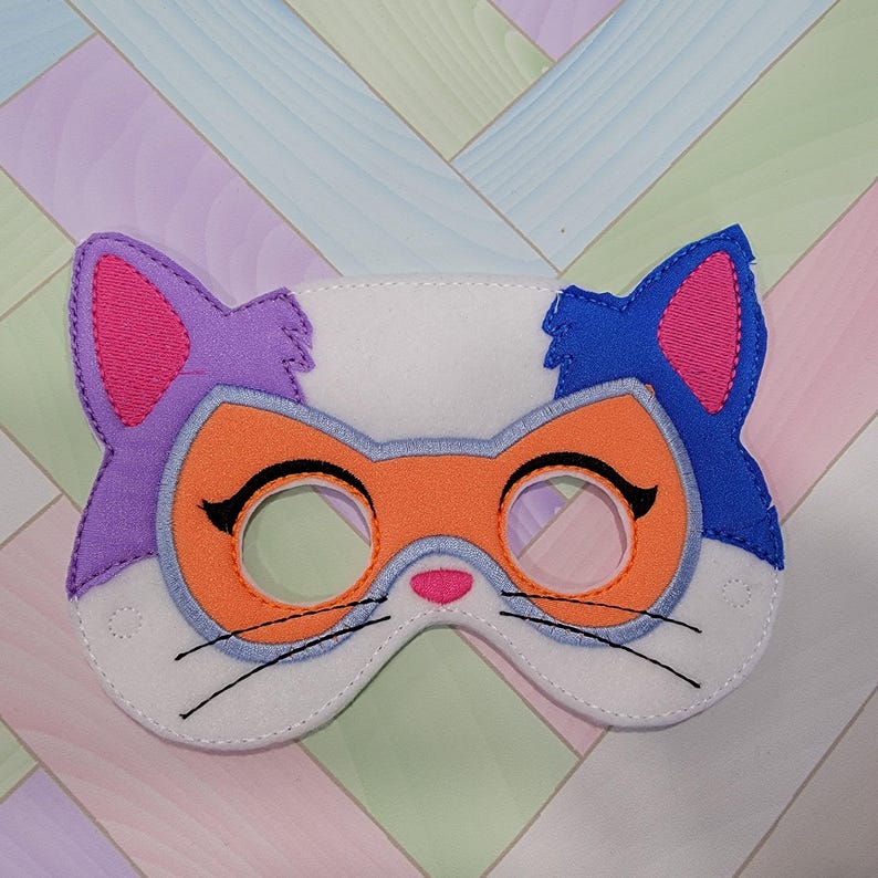 May include: A felt cat mask featuring a white base with a purple left ear and a blue right ear. The mask includes an orange eye patch area, pink inner ears, a small pink nose, and black embroidered eyelashes and whiskers around the eye openings.