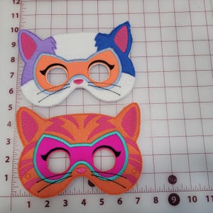 May include: Two felt cat masks positioned on a cutting mat with centimetre markings. The top mask is white with a purple ear, blue ear, and orange eye area. The bottom mask is orange with pink stripes and a bright pink eye area with teal trim.