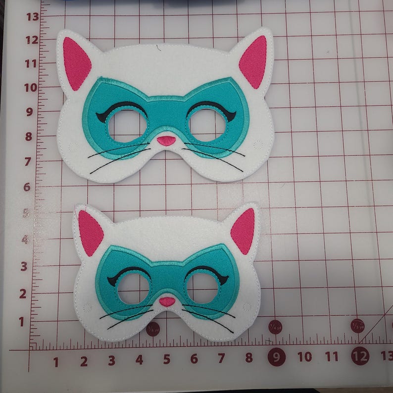 May include: Two white felt cat masks with pink inner ears, a teal eye mask design, black embroidered eyelashes, and whiskers. The masks are positioned on a cutting mat with metric measurements, showing the larger mask is 15 cm wide.