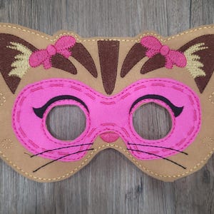 May include: A tan felt cat mask featuring a bright pink eye area with black embroidered eyelashes and whiskers. The mask includes dark brown ear details, light yellow inner ear accents, and two small pink bows stitched above the eye openings.