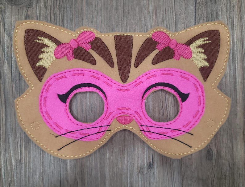 May include: A tan felt cat mask featuring a bright pink eye area with black embroidered eyelashes and whiskers. The mask includes dark brown ear details, light yellow inner ear accents, and two small pink bows stitched above the eye openings.