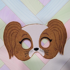May include: A brown and white felt mask shaped like a dog face with long floppy ears. The mask features embroidered black eyes with eyelashes, a small black nose, and whiskers. The white section extends from the forehead to the nose area.