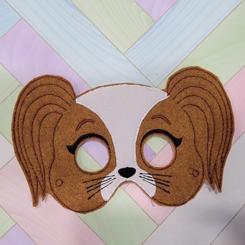 May include: A brown and white felt mask shaped like a dog face with long floppy ears. The mask features embroidered black eyes with eyelashes, a small black nose, and whiskers. The white section extends from the forehead to the nose area.