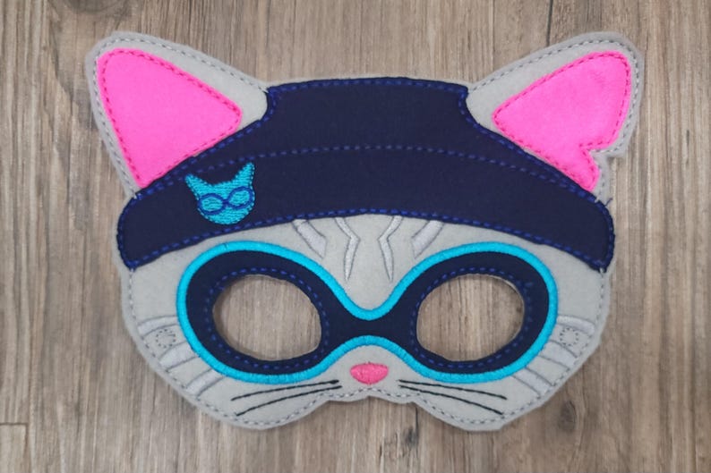 May include: A grey felt cat mask featuring bright pink inner ears, a dark navy blue beanie cap with a small light blue cat icon, and dark navy blue eye patches outlined in light blue. The mask includes a small pink nose and black stitched whiskers.